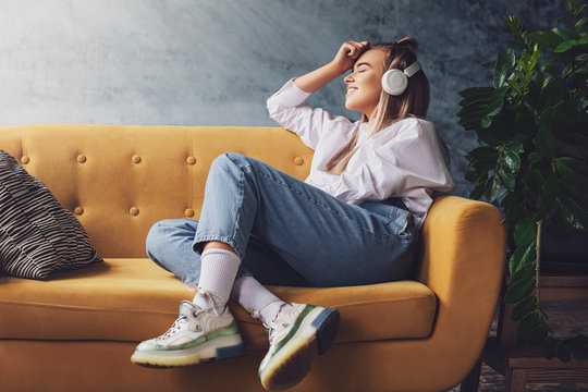 Beautiful Blonde Woman In White Headphones Sits On A Yellow Sofa And Enjoys Listening To Music, Podcast.