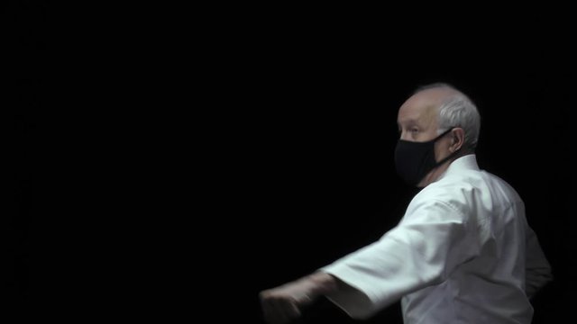In a black medical mask in a karate stance, an old athlete trains blocks and punches arms