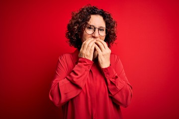 Middle age beautiful curly hair woman wearing casual shirt and glasses over red background laughing and embarrassed giggle covering mouth with hands, gossip and scandal concept