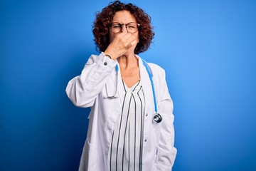 Middle age curly hair doctor woman wearing coat and stethoscope over blue background smelling something stinky and disgusting, intolerable smell, holding breath with fingers on nose. Bad smell