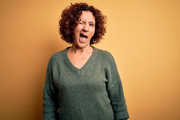 Middle age beautiful curly hair woman wearing casual sweater over isolated yellow background winking looking at the camera with sexy expression, cheerful and happy face.
