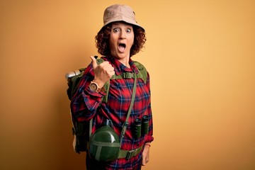 Middle age curly hair hiker woman hiking wearing backpack and water canteen using binoculars Surprised pointing with hand finger to the side, open mouth amazed expression.