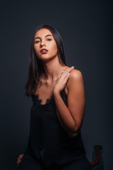 Sexy brunette wearing a black tank top and black pants posing while sitting on the top edge of a chair inside a studio on a dark background