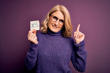 Middle age beautiful blonde woman wearing glasses holding reminder paper with yes message surprised...