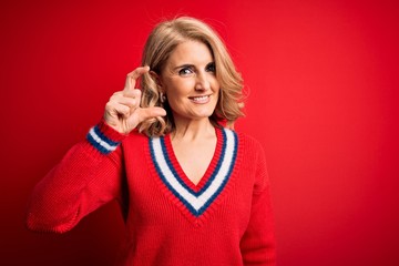 Middle age beautiful blonde woman wearing casual sweater over isolated red background smiling and confident gesturing with hand doing small size sign with fingers looking and the camera. Measure 
