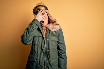 Middle age beautiful blonde skier woman wearing snow sportwear and ski goggles peeking in shock covering face and eyes with hand, looking through fingers with embarrassed expression.