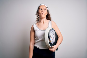 Young beautiful woman with curly hair holding weighing machine over white background making fish face with lips, crazy and comical gesture. Funny expression.