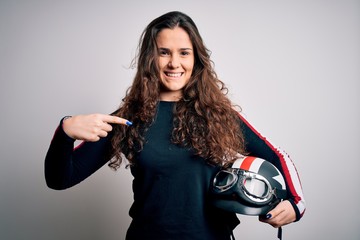 Obraz premium Beautiful motorcyclist woman with curly hair holding moto helmet over white background with surprise face pointing finger to himself