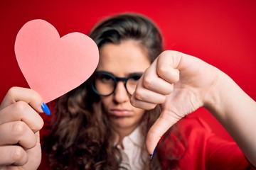 Young beautiful woman with curly hair holding paper heart over isolated red background with angry...