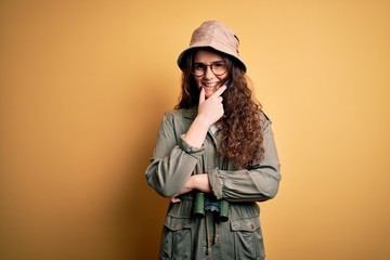 Young beautiful tourist woman on vacation wearing explorer hat and binoculars looking confident at the camera smiling with crossed arms and hand raised on chin. Thinking positive.