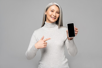 Naklejka premium happy asian woman in turtleneck with grey hair pointing at smartphone with blank screen isolated on grey
