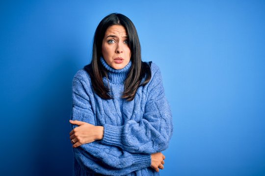 Young brunette woman with blue eyes wearing casual turtleneck sweater shaking and freezing for winter cold with sad and shock expression on face