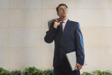 Businessman in formal clothes carrying office bag in one hand and a laptop in other looking away
