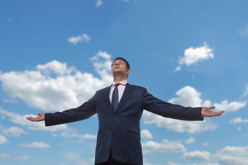 Businessman in formal clothes standing with open arms and closed eyes against a cloudy blue sky
