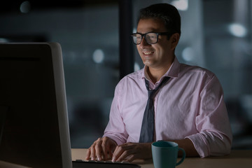 Smiling businessman in eyeglasses working on computer sitting late in office with a cup of coffee...