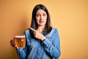 Young woman with blue eyes drinking jar of beer standing over isolated yellow background Pointing...