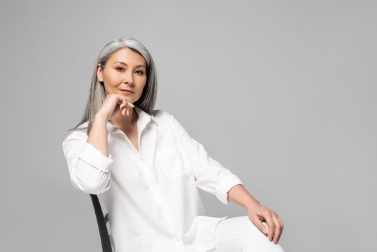 Serious Asian Woman With Grey Hair Sitting On Chair Isolated On Grey