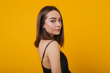 Obraz premium Pleasant young lady looking over shoulder while posing on yellow background. Studio portrait of lovely girl in black tank-top chilling during photoshoot.