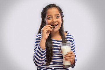 Smiling girl eating a cream biscuit while holding a glass of milk in one hand
