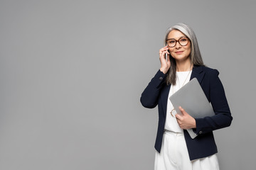 corporate asian businesswoman with grey hair talking on smartphone and holding laptop isolated on grey