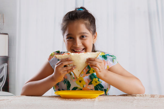 Smiling girl eating a bread sandwich sitting at dining table
