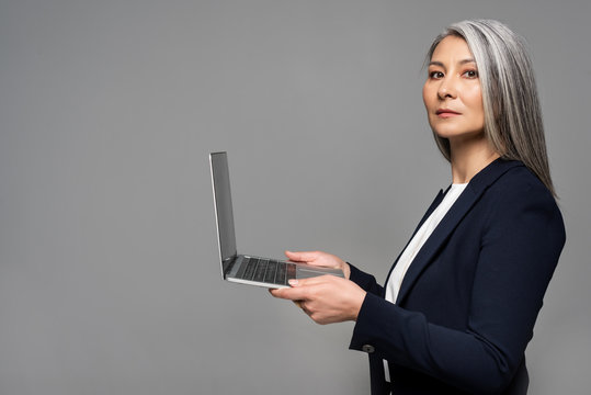 Attractive Serious Asian Businesswoman With Grey Hair Using Laptop Isolated On Grey