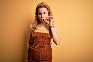 Young beautiful blonde woman wearing overalls and diadem standing over yellow background mouth and lips shut as zip with fingers. Secret and silent, taboo talking