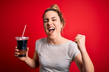Beautiful blonde woman drinking cola fizzy beverage to refreshment over red background screaming proud and celebrating victory and success very excited, cheering emotion
