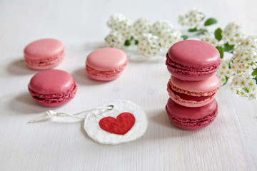 Pink macarons / macaroon on a white wooden table with tender white blossoms in the background and a gift tag with a heart.