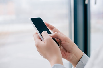 cropped view of professional businesswoman using smartphone with blank screen near window in office