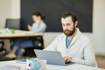 Handsome young man with beard on face sitting at office table working on his laptop with serious facial expression