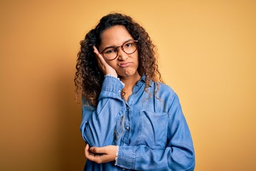 Beautiful woman with curly hair wearing casual denim shirt and glasses over yellow background thinking looking tired and bored with depression problems with crossed arms.