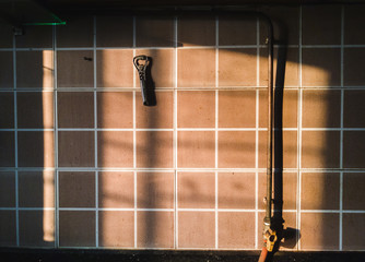Evening light falls on a tiled wall with a bottle opener and a cooking gas pipeline with a knob  inside a kitchen.