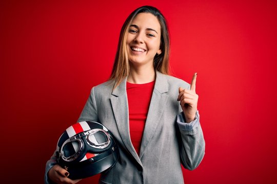 Young beautiful blonde motorcyclist woman holding motorcycle helmet over red background surprised with an idea or question pointing finger with happy face, number one