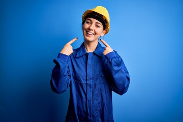 Young beautiful worker woman with blue eyes wearing security helmet and uniform smiling cheerful showing and pointing with fingers teeth and mouth. Dental health concept.