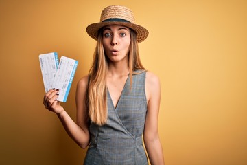 Young blonde tourist woman with blue eyes on vacation wearing hat holding boarding pass scared in shock with a surprise face, afraid and excited with fear expression