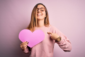 Young beautiful blonde romantic woman wearing glasses holding big pink heart with surprise face pointing finger to himself