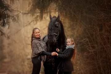 Mother and daughter  with Black Beautiful  Friesian stallion with long hair outdoor portrait in an autumn forest