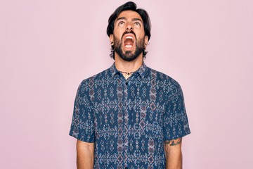 Young handsome hispanic bohemian man wearing hippie style over pink background angry and mad screaming frustrated and furious, shouting with anger. Rage and aggressive concept.
