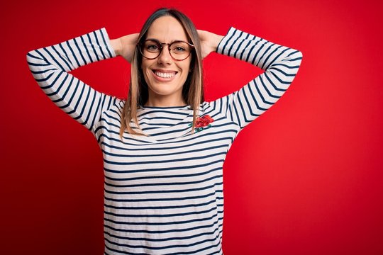 Young beautiful blonde woman with blue eyes wearing glasses standing over red background relaxing and stretching, arms and hands behind head and neck smiling happy