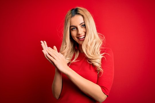 Young beautiful blonde woman wearing casual t-shirt standing over isolated red background clapping and applauding happy and joyful, smiling proud hands together