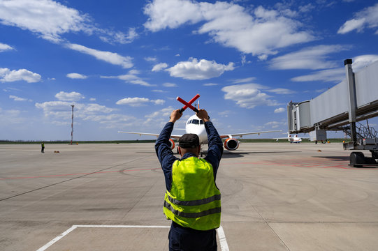 A male airport marshal with wands in a yellow uniform helps park the plane. The supervisor meets a passenger plane at the airport. The plane steers into the parking lot.