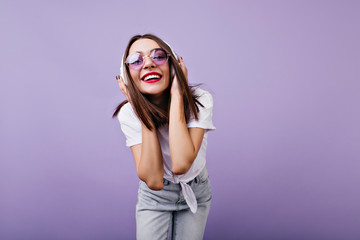 Glad girl in sunglasses touching her headphones with smile. Indoor photo of gorgeous female model in jeans isolated on purple background.