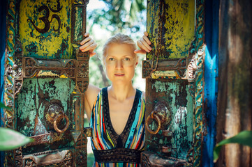 Beautiful blonde opening the doors of vintage gate. Portrait of young woman in authentic style against a half-open old gates. Concept of discovery, new life, adventure. Om Symbol on the door.