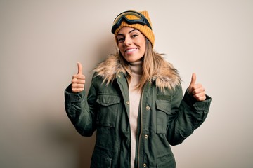 Young brunette skier woman wearing snow clothes and ski goggles over white background success sign doing positive gesture with hand, thumbs up smiling and happy. Cheerful expression and winner gesture