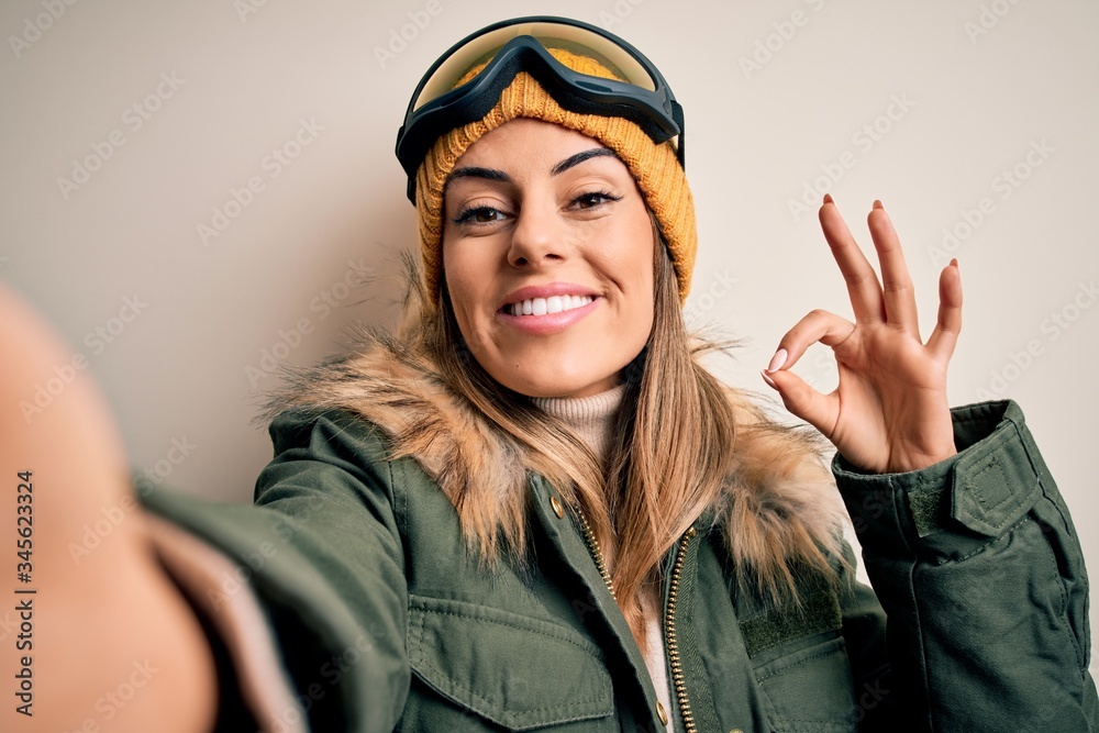 Sticker Young brunette skier woman wearing snow clothes and ski goggles make selfie by camera doing ok sign with fingers, excellent symbol