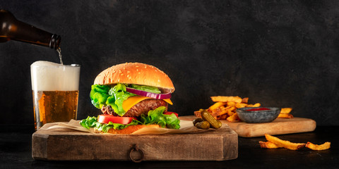Burger and beer panorama. Hamburger with beef, cheese, onion, tomato, and green salad, a side view on a dark background with a place for text. Selective focus