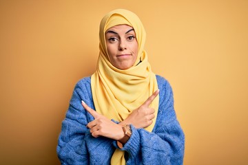 Young beautiful brunette muslim woman wearing arab hijab over isolated yellow background Pointing to both sides with fingers, different direction disagree