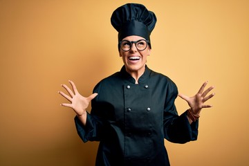 Young beautiful brunette chef woman wearing cooker uniform and hat over yellow background crazy and mad shouting and yelling with aggressive expression and arms raised. Frustration concept.