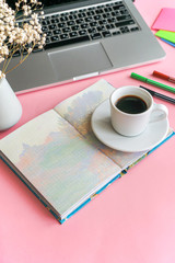 a coffee mug stands on a notebook, next to it lies a phone, pens and a laptop